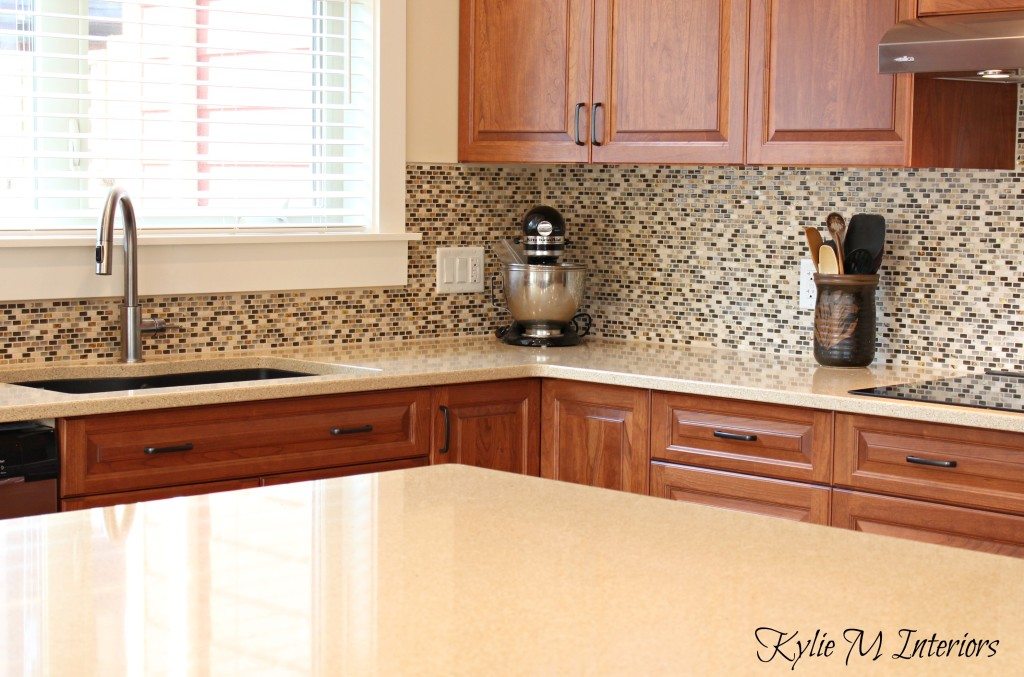 cream quartz countertops, cherry kitchen cabinets, small mosaic tile backsplash, cable knit paint colour, black hardware and black appliance with an island