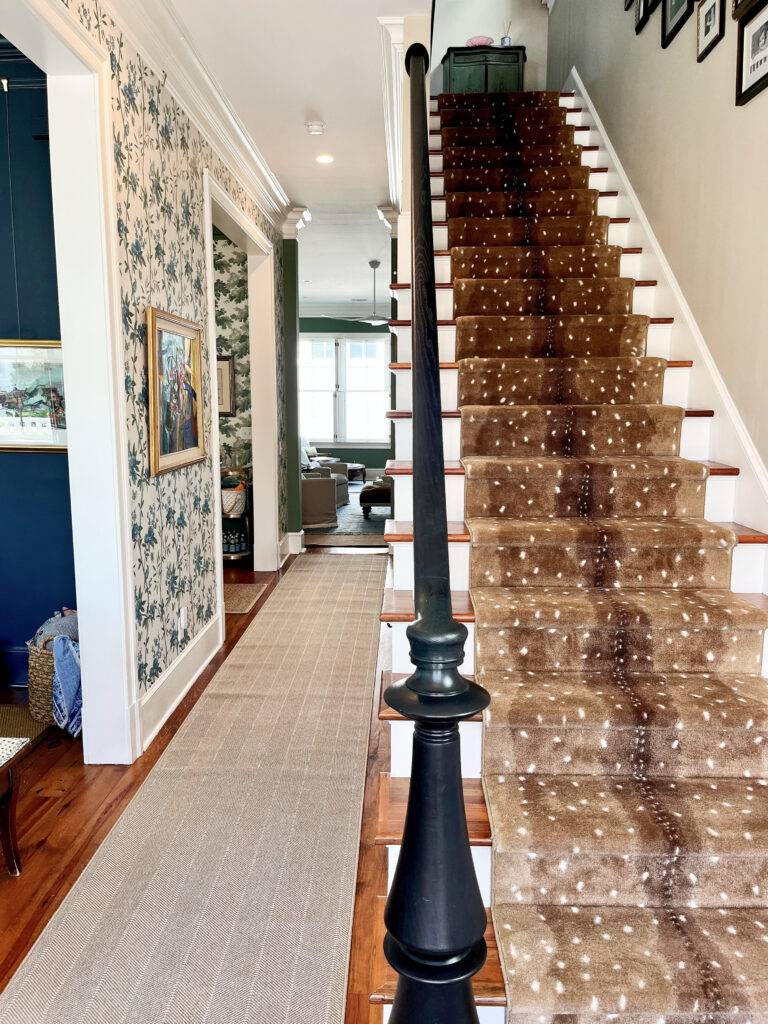 Foyer or entryway with deerskin pattern stair runner, wood steps, black hand railing, wallpaper, beige striped runner on oak floor, white trim.