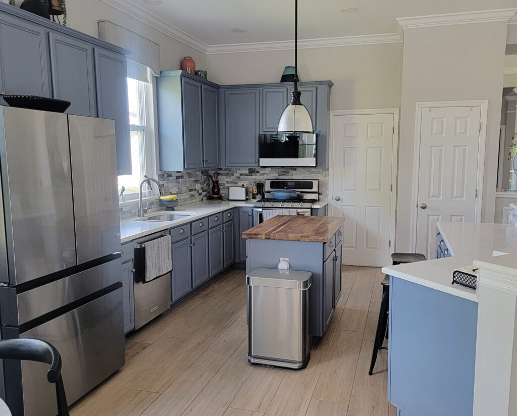 Blue paint color on painted kitchen cabinets and island, butcher block and white quartz, stacked marble look stone backsplash, light oak wood flooring, Sherwin Williams On the Rocks walls