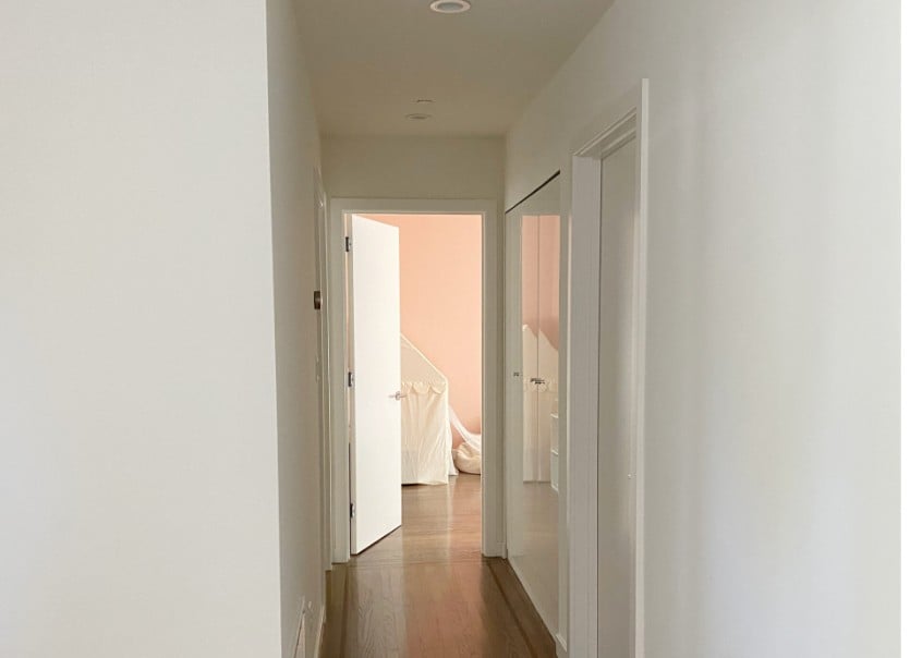 Kelly Moore Swiss Coffee warm white paint color with medium stained oak flooring down darker hallway, pink bedroom at end