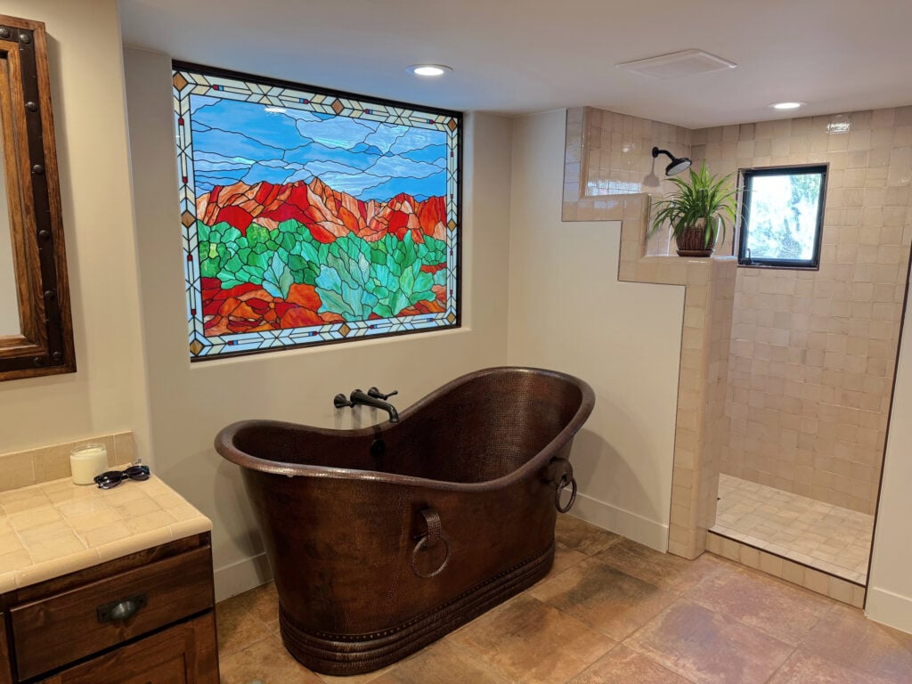 Stained glass window in bathroom with freestanding copper bathtub, beige tile floor and walk in shower, wood vanity