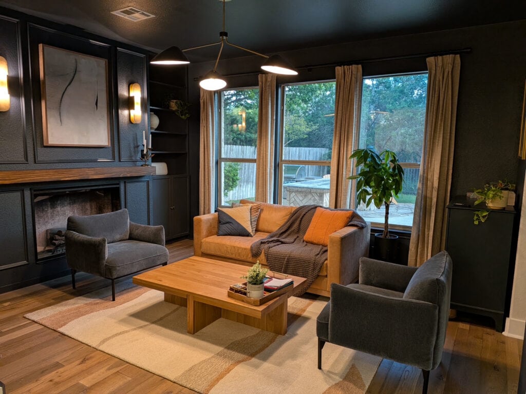 Moody den or sitting room, wood floor, Sherwin Williams Cybersepace dark gray-blue walls, built-ins and fireplace with wood mantel. Wall sconces, warm sofa, and dark gray-blue accent chairs