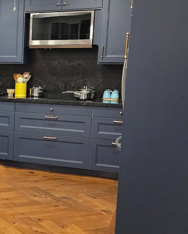 Benjamin Moore Hale Navy, navy blue painted kitchen cabinets with black granite countertop and slab backsplash. No natural light, herringbone wood flooring