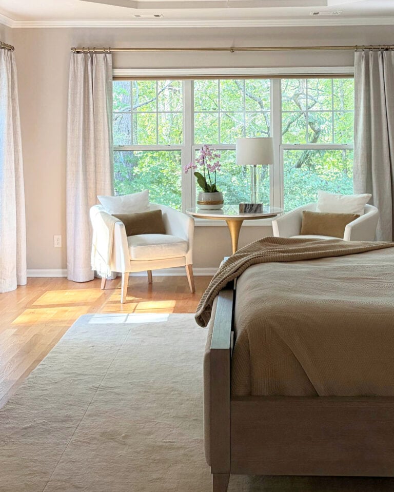 Primary, northwest facing bedroom painted Sherwin Williams Popular Gray, accent chairs by window with drapes and wood floor. Pure White trim and tray ceilng